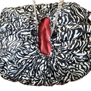 May include: A round, drawstring cosmetic bag featuring a black and white zebra print pattern on the exterior. The interior of the bag is lined with a solid red fabric. Two thin, light-coloured cords are attached to the top for cinching the bag closed.