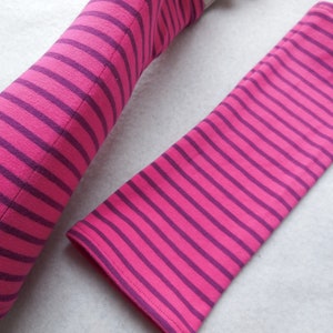 May include: Pink and purple striped arm warmers. The arm warmers are made of a soft fabric and feature evenly spaced horizontal stripes. The stripes alternate between a bright pink and a deep purple color. The arm warmers are shown on a white surface.