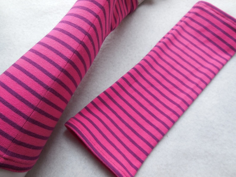 May include: Pink and purple striped arm warmers. The arm warmers are made of a soft fabric and feature evenly spaced horizontal stripes. The stripes alternate between a bright pink and a deep purple color. The arm warmers are shown on a white surface.
