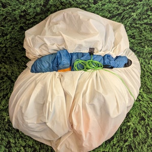 May include: A white, drawstring stuff sack with a blue and orange tent packed inside. The tent is partially visible and has a green cord attached to it.