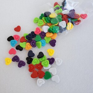 May include: A collection of colorful heart-shaped embellishments in various shades, including red, pink, yellow, green, purple, black, and white. Some hearts are solid, while others have a heart outline. The embellishments are scattered on a white surface.