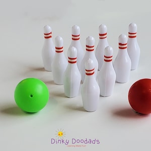 Puede incluir: Un juego de bolos en miniatura con bolos blancos con rayas rojas en la parte superior, dispuestos en un triángulo. También hay una bola verde con un agujero y una bola roja. El texto "Dinky Doodads" está en la parte inferior.