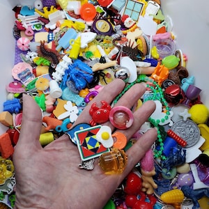 May include: A hand displaying a selection of colourful charms and trinkets, such as a red crab, a pink ring, and a square charm with a geometric design. The background is filled with various small objects in diverse shapes and colours.
