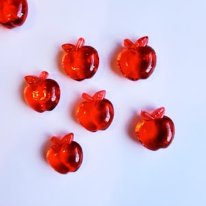 May include: Several translucent red apple-shaped beads with stems and leaves are scattered on a white background. The beads, possibly glass, have a glossy finish. The arrangement is decorative and could be used for crafts or embellishments.