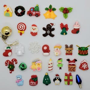 Puede incluir: Una colección de pequeños dijes planos de resina con temática navideña que presentan varias formas, incluyendo una casa, una corona, un camión, campanas, acebo, dulces, Santa Claus, un hombre de jengibre, un calcetín, un copo de nieve y un tren.