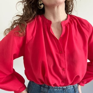 May include: A bright red, long-sleeved blouse with a button-up front and a gathered neckline. The blouse has a relaxed fit and is made of a lightweight fabric.