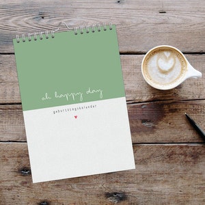 Könnte beinhalten: Ein grüner und weißer Spiral-Kalender mit dem Text "oh happy day" und "Geburtstagskalender" auf dem Cover. Der Kalender liegt auf einer Holzoberfläche mit einer Tasse Kaffee und einem Stift.