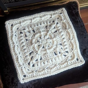 Könnte beinhalten: Ein weißes quadratisches Kissen aus Häkelarbeit mit einem Blumenmuster. Das Kissen ist mit einem filigranen Stichmuster gearbeitet und hat eine weiche, strukturierte Oberfläche.