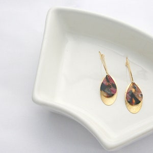 May include: A pair of gold dangle earrings with a teardrop-shaped design. The teardrop is made of a layered material with a gold base and a top layer of brown, black, and red marbled resin.