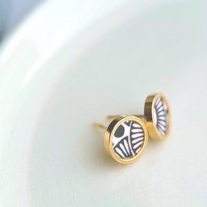 Peut inclure: Boucles d'oreilles à clous dorées avec un motif géométrique noir et blanc. Les boucles d'oreilles sont rondes et présentent un style simple et élégant. Les boucles d'oreilles sont présentées sur une surface blanche.