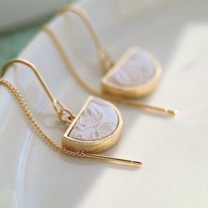 May include: A pair of gold-tone threader earrings featuring semi-circular pendants. Each pendant has a textured gold metal rim and a white, embossed inlay with a delicate, repeating floral or shell-like pattern on the surface.