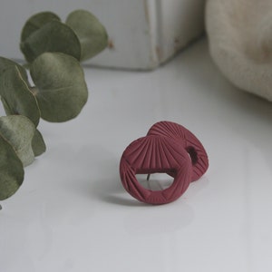 May include: A pair of burgundy clay earrings with a geometric design. The earrings are round and have a textured surface.