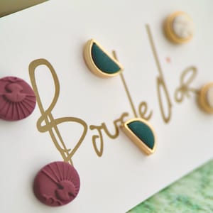 Könnte beinhalten: Eine Auswahl an Ohrsteckern auf einer weißen Karte mit goldener Schrift. Die Ohrringe umfassen goldgerahmte teal Halbmonde, kleine beige Kreise und maroon runde Ohrstecker mit einem Sonnenaufgangsdesign.