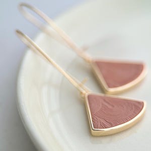 Peut inclure: Une paire de boucles d'oreilles dorées avec un insert triangulaire en émail rose. Les boucles d'oreilles ont un design délicat et minimaliste, avec un motif tourbillonnant sur l'émail. Les boucles d'oreilles sont présentées sur une surface blanche.