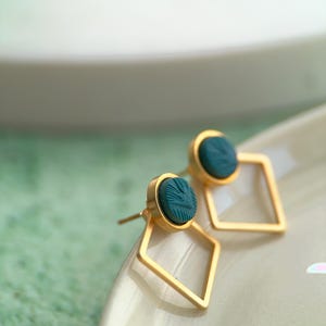 Peut inclure: Une paire de boucles d'oreilles dorées avec un centre circulaire turquoise et un cadre en forme de losange. Le centre turquoise présente un motif texturé. Les boucles d'oreilles sont présentées sur une surface blanche et verte.