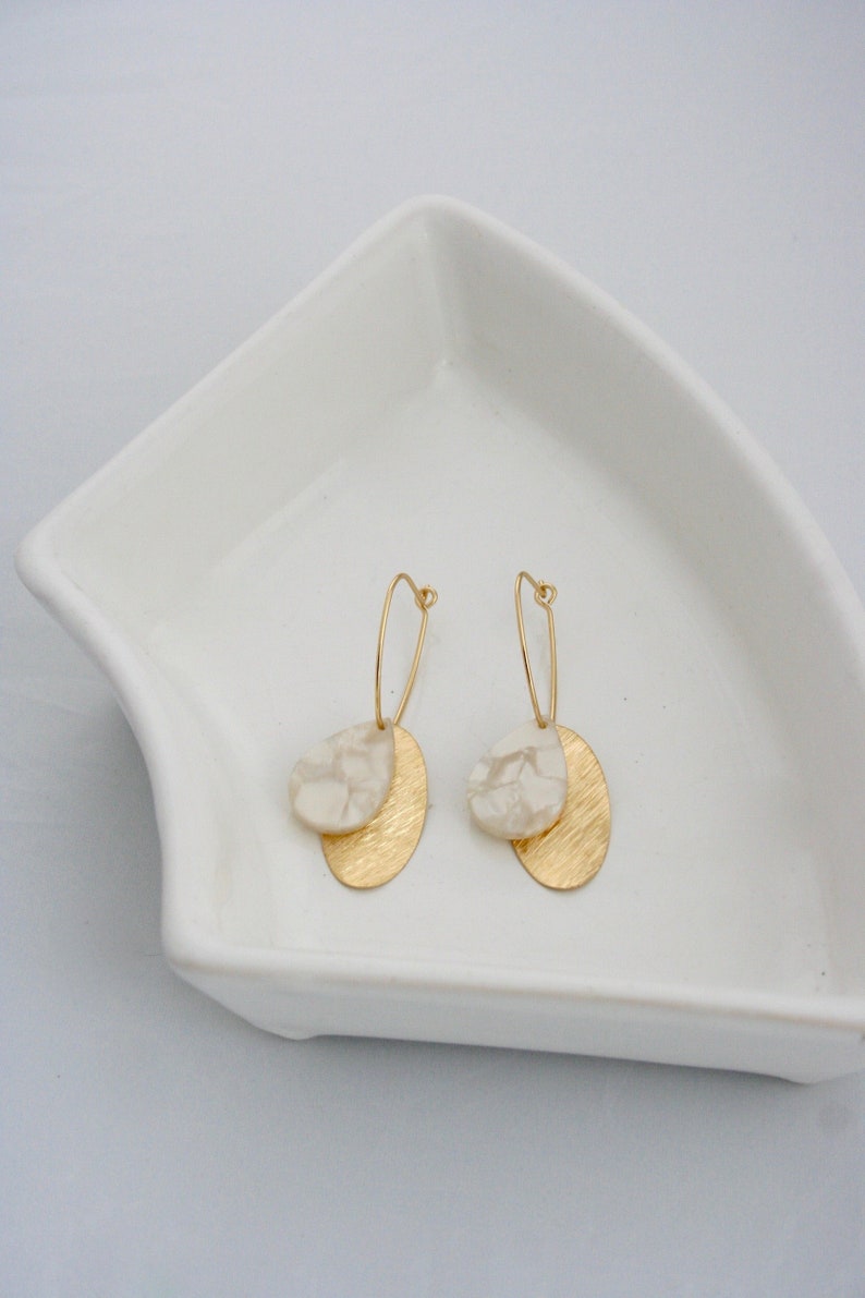 May include: A pair of gold hoop earrings with white marble-like oval charms hanging from them. The earrings are displayed on a white ceramic dish.