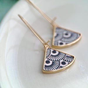 Puede incluir: Un par de pendientes dorados. Cada pendiente presenta un colgante en forma de abanico con un patrón geométrico en blanco y negro. Los pendientes se muestran sobre una superficie blanca.