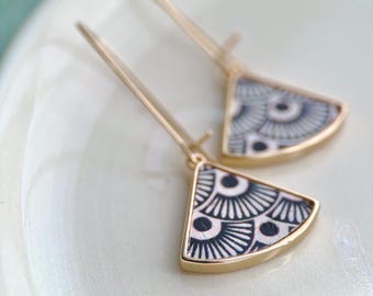 May include: A pair of gold-toned drop earrings featuring a fan-shaped pendant. Each pendant has a flat, triangular base with a rounded edge, decorated with a black and white geometric pattern of repeating semicircles and radiating lines.