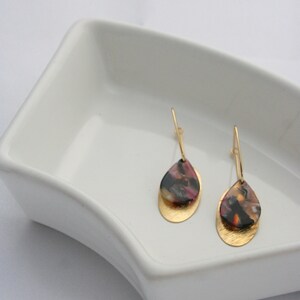 May include: A pair of gold dangle earrings with a teardrop-shaped, multi-coloured resin stone. The resin stone has shades of pink, black, and gold.