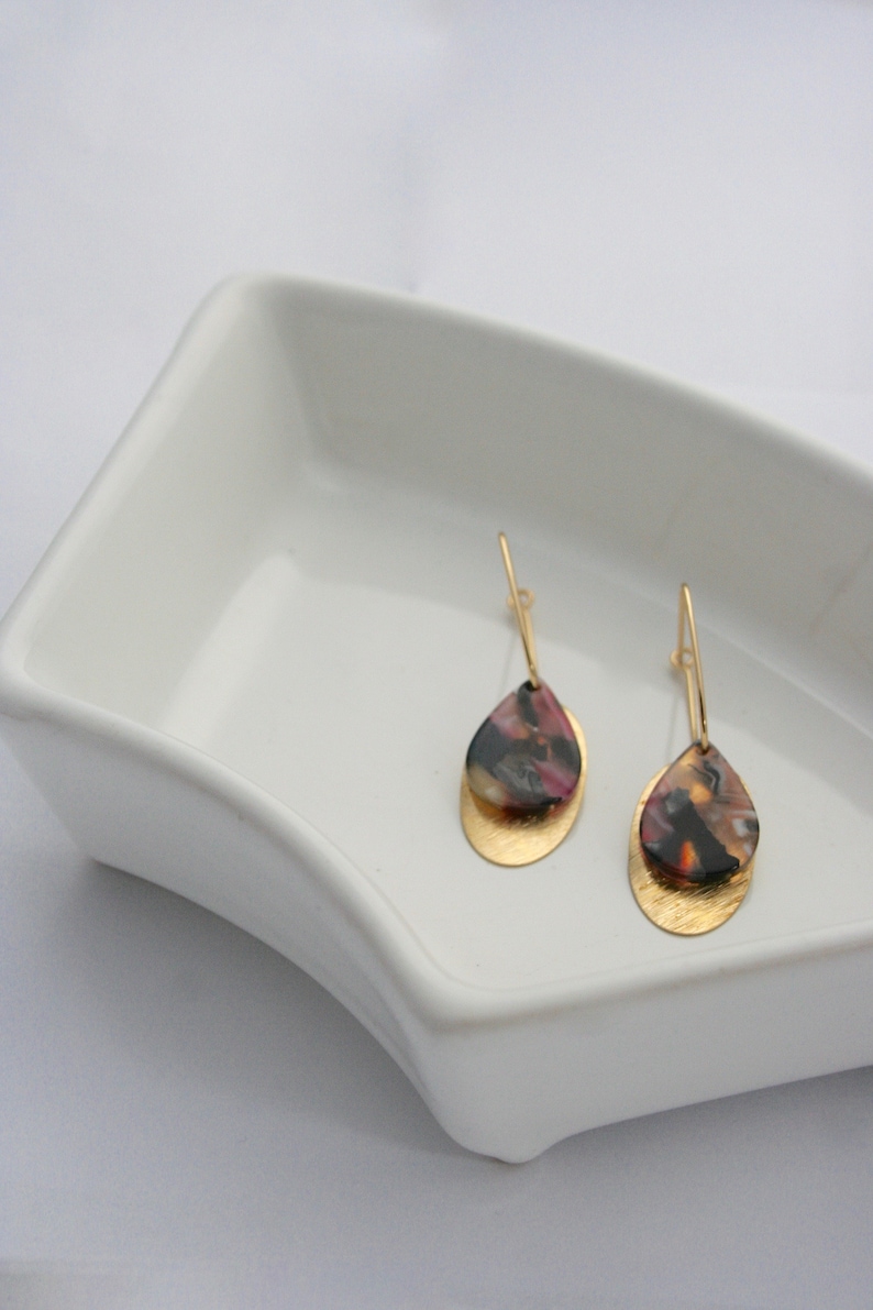May include: A pair of gold dangle earrings with a teardrop-shaped, multi-coloured resin stone. The resin stone has shades of pink, black, and gold.