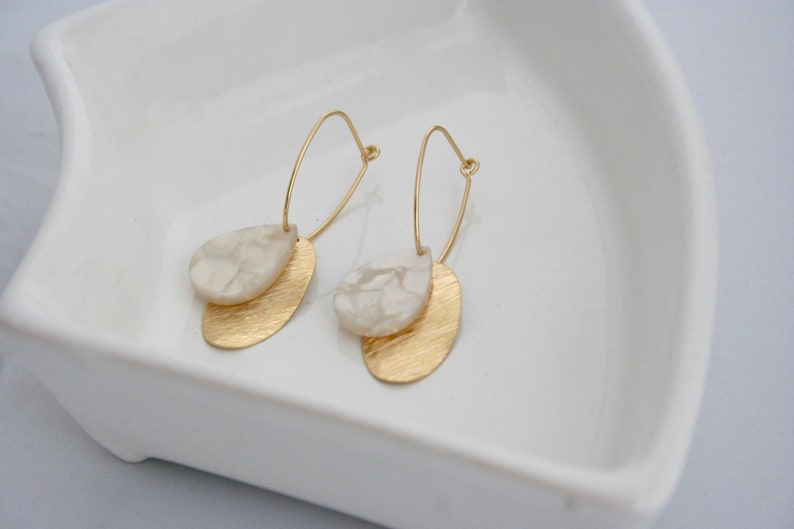 May include: A pair of gold hoop earrings with a white, marbled, teardrop-shaped pendant hanging from each hoop.