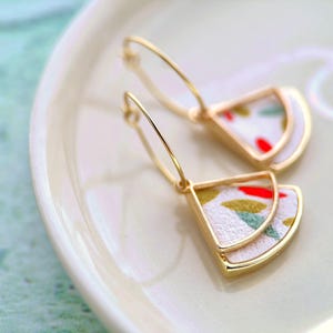 May include: Gold-coloured hoop earrings with a distinctive fan-shaped design. The earrings have a white fabric inlay with red, green, and yellow accents. The earrings are presented on a white ceramic plate.