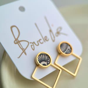 Puede incluir: Pendientes dorados con un diseño geométrico. Cada pendiente presenta un perno circular con un patrón en blanco y negro, conectado a un marco cuadrado. Los pendientes se exhiben en una etiqueta blanca con la inscripción "Boucle de" en oro.