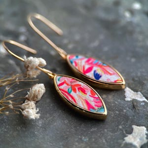 Könnte beinhalten: Ein Paar goldene Ohrhänger mit rosa und weißen floralen Stoffakzenten. Die Ohrringe haben eine Tropfenform und sind mit Gold verziert.