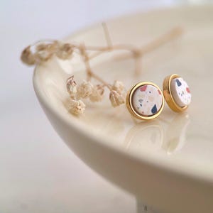 May include: A pair of gold stud earrings with white, pink and blue terrazzo-style designs.