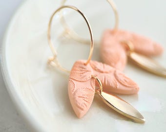 May include: A pair of hoop earrings featuring thin gold-toned metal hoops. Each earring is adorned with a peach-coloured, textured leaf-shaped charm and a smooth, polished gold-toned metal leaf-shaped charm hanging from the bottom of the hoop.
