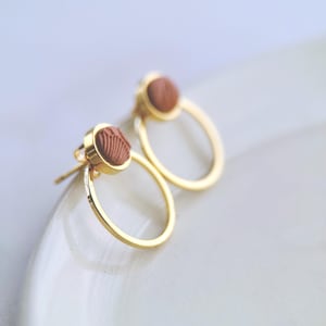 May include: A pair of gold-coloured hoop earrings featuring a circular, brown, textured design. The earrings have a post-back closure. The earrings are displayed on a white surface.