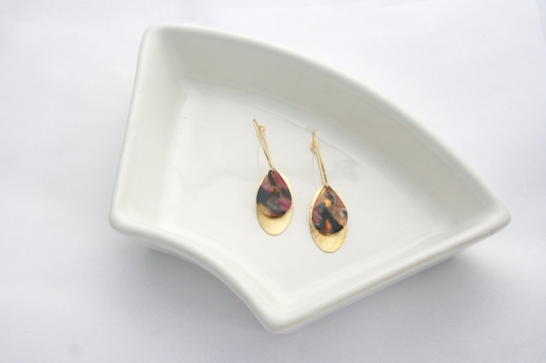 May include: A pair of gold dangle earrings with a teardrop-shaped design. The teardrop is made of a layered material with a gold base and a top layer of brown, black, and red marbled resin.