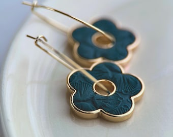 Puede incluir: Un par de pendientes de aro dorados con colgantes en forma de flor de color verde oscuro. Los colgantes tienen una superficie texturizada y un borde dorado. Los pendientes se muestran sobre una superficie blanca.