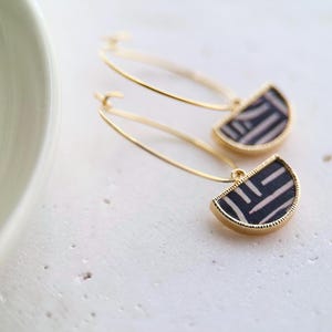 Könnte beinhalten: Goldene Creolen mit einem halbkreisförmigen Anhänger. Der Anhänger hat ein schwarz-weißes geometrisches Muster und einen goldenen Rand. Die Ohrringe werden auf einer weißen Oberfläche präsentiert.