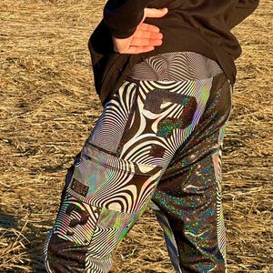 May include: Black and white iridescent pants with a swirling pattern and a black pocket on the left leg. The pants have a reflective, glittery finish.