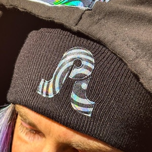 May include: A black beanie with a holographic, striped letter 'A' logo. The beanie is made of a soft, knit fabric.
