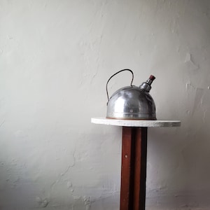 May include: A silver kettle with a red knob on a white table with a brown wooden base.