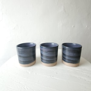 May include: Three ceramic cups with a dark blue, horizontally striped design. The cups have a light tan base and a smooth, rounded shape. They are arranged on a white, round surface.