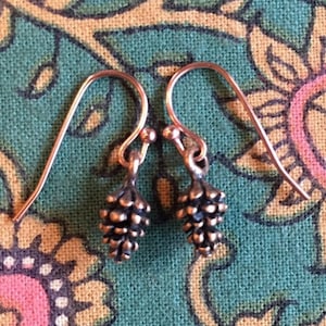 May include: A pair of small, copper-colored earrings with a pine cone design. The earrings are hanging from a floral patterned fabric.
