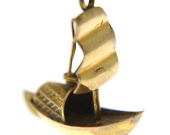 May include: A gold-colored charm in the shape of a sailboat with a single sail. The sail is detailed with ridges and the boat has a small, detailed hull.