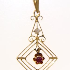 May include: A gold-colored pendant with a geometric design. The pendant features a small, red flower-shaped gemstone at the center and a small, light-colored gemstone above it. The pendant is suspended from a gold bail.