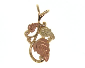 May include: A gold-tone pendant featuring a vine design with three leaves in tri-color gold: yellow, rose, and green. The pendant has a loop at the top for a chain and small grape clusters.