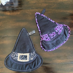 May include: Two black witch hat shaped oven mitts. One mitt has a purple and black leopard print trim. The other mitt has a black and white spiderweb design.