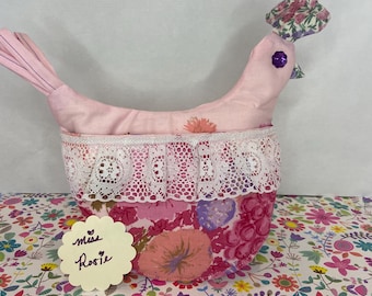 May include: A pink fabric chicken with a floral print and lace trim. The chicken has a purple button for an eye and a floral fabric bow on its head. The chicken is sitting on a floral patterned surface with a yellow note that says "Miss Rosie".