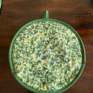 Peut inclure: Une manivelle de pot en tissu vert et blanc avec un motif de feuilles. La manivelle de pot a une bordure en tissu vert et une poignée en boucle.