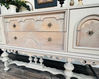 Puede incluir: Un aparador antiguo en tonos gris claro y madera, con tallas ornamentadas y herrajes de latón. El aparador tiene dos cajones y una puerta de armario. Candelabros decorativos y un cuadro enmarcado encima.