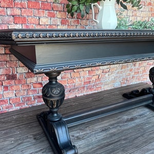 May include: A black, ornate table with a decorative edge and carved legs. The table has a dark finish with gold accents. A white pitcher with flowers sits on the table's surface, against a brick wall background.