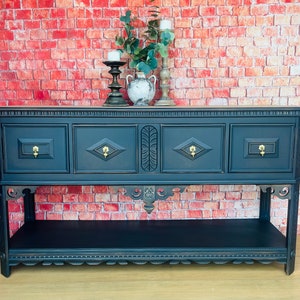 May include: A dark blue console table with gold-colored drawer pulls and decorative carvings. The table has a lower shelf and is set against a red brick wall. Decorative items, including candles and a vase, sit on top.