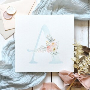 May include: A watercolor painting of the letter A with a floral design in shades of pink, white, and green. The letter is painted on a light blue background.