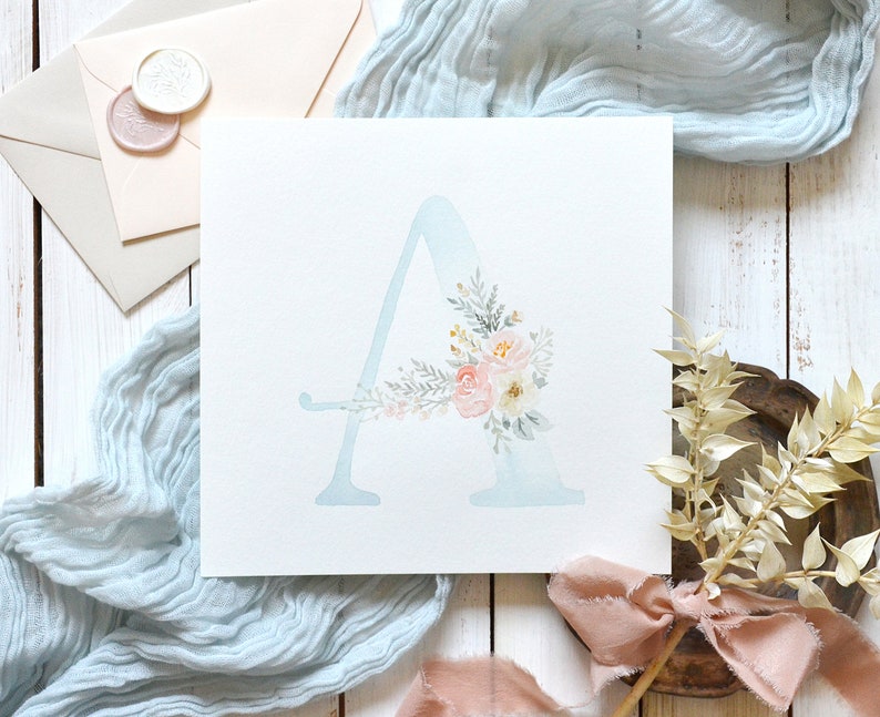 May include: A watercolor painting of the letter A with a floral design in shades of pink, white, and green. The letter is painted on a light blue background.